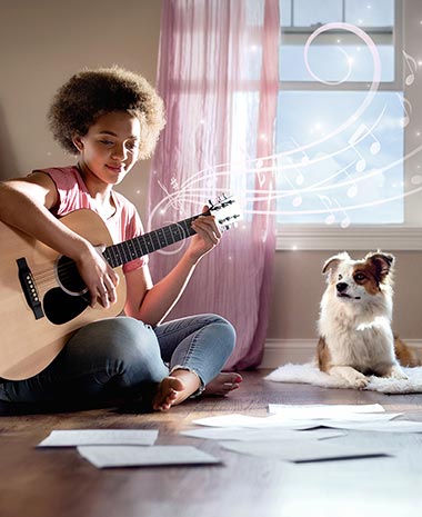 Girl playing guitair with her dog sitting next to her.